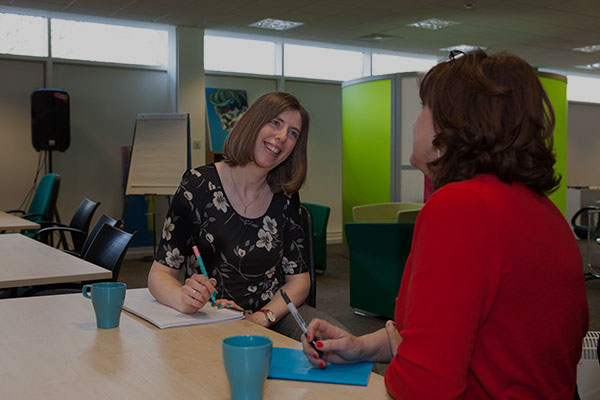 Two women sit opposite each other at a table, they are having a conversation and both have notepads and pens in front of them