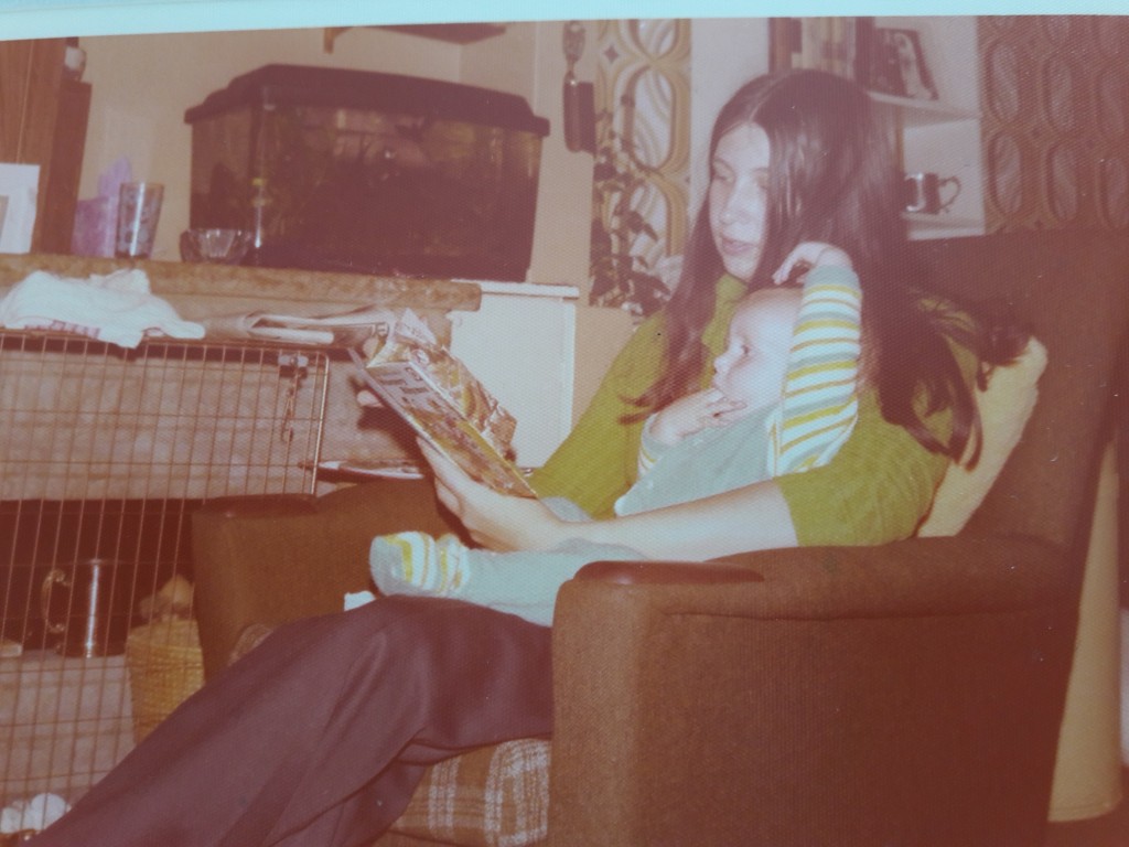 Sarah as a baby, sitting on a sofa with her Mum reading a book to her