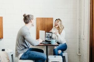 A young woman and a man sitting at a table in a casual-looking office having a conversation and laughing