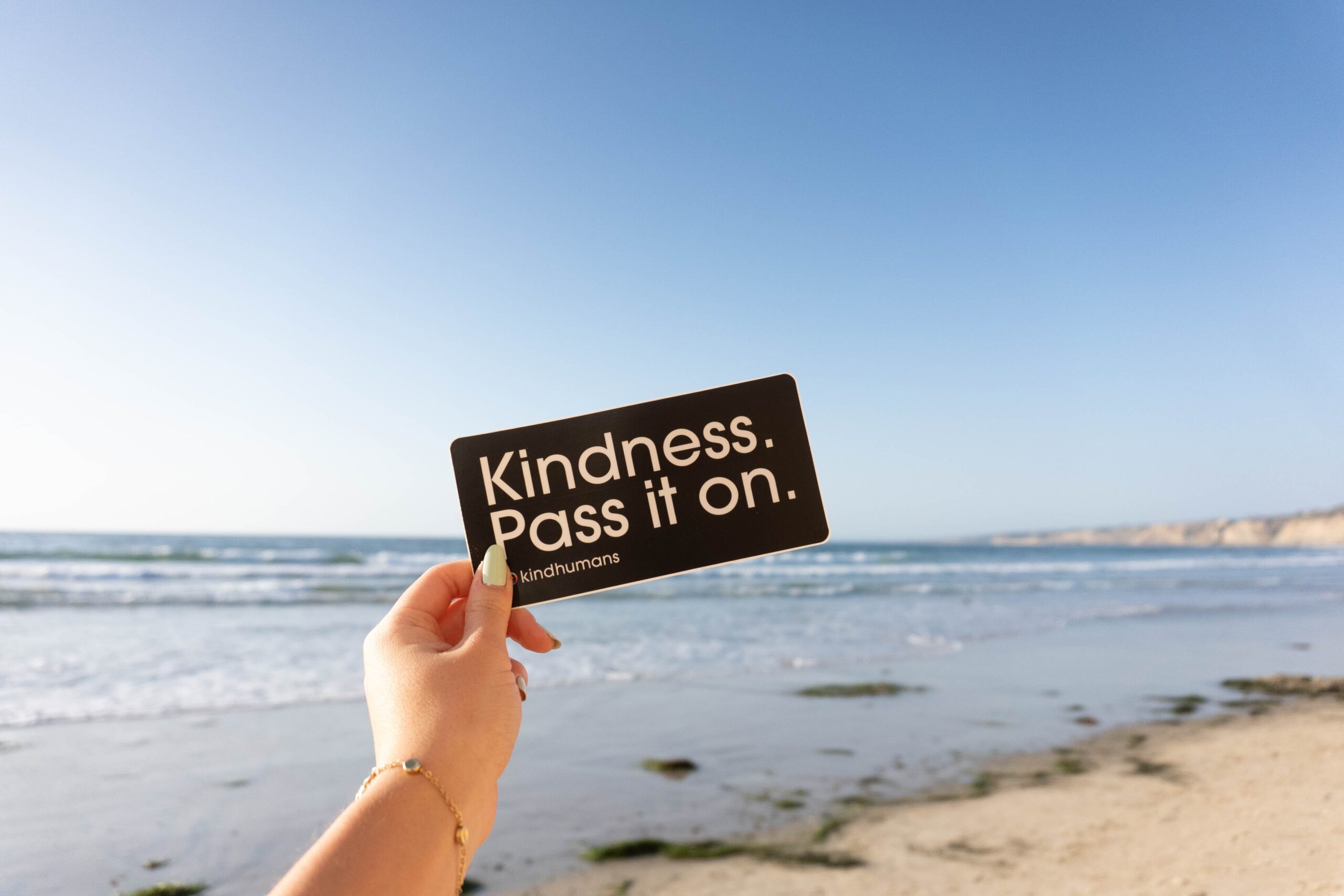 A photo of a hand holding a note that says Kindness, pass it on. Behind the note we can see blue sea and blue sky and a little bit of beach