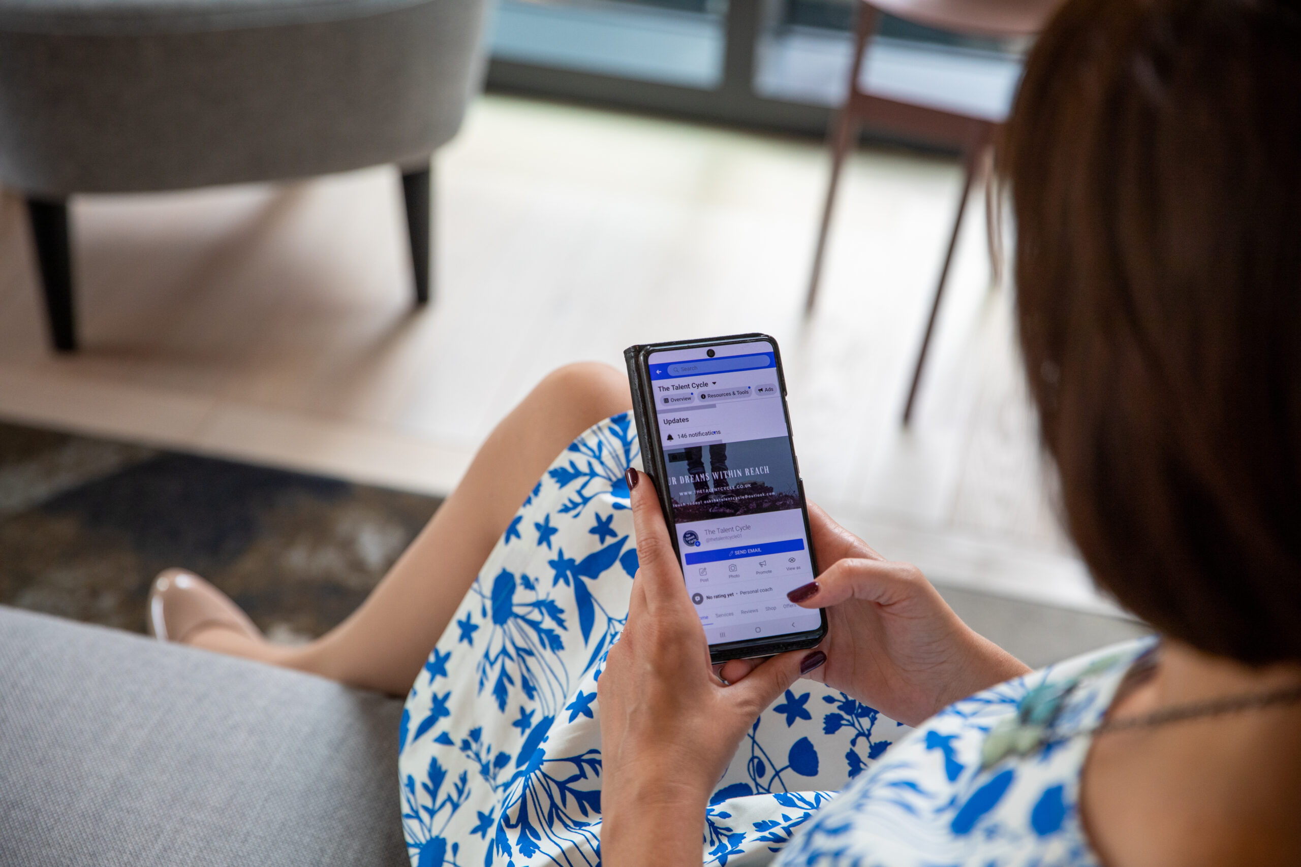 Caroline sits on a grey sofa in an office wearing a smart blue and white dress and looking at a career page on her smartphone