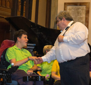 A member of the Choir wearing a green shirt and sitting in a wheelchair shakes hands with a man who is wearing a white shirt and standing in front of him