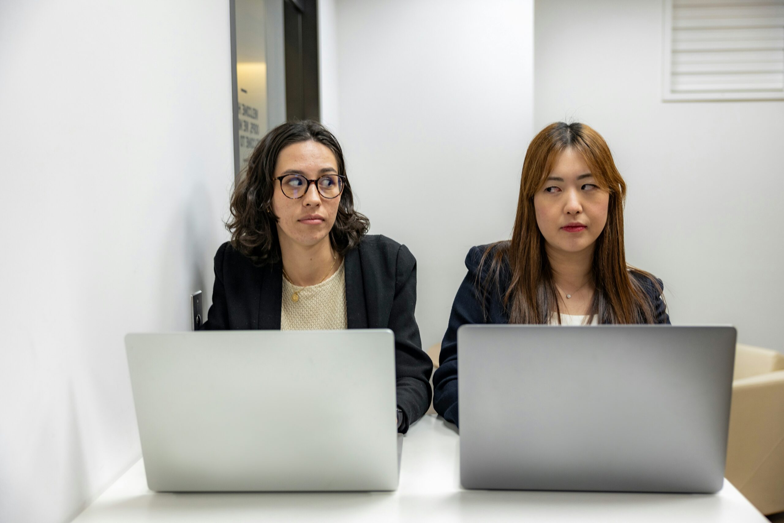 Two women sitting next to each other with laptops in front of them - they are looking at each with disgruntled looks on their faces